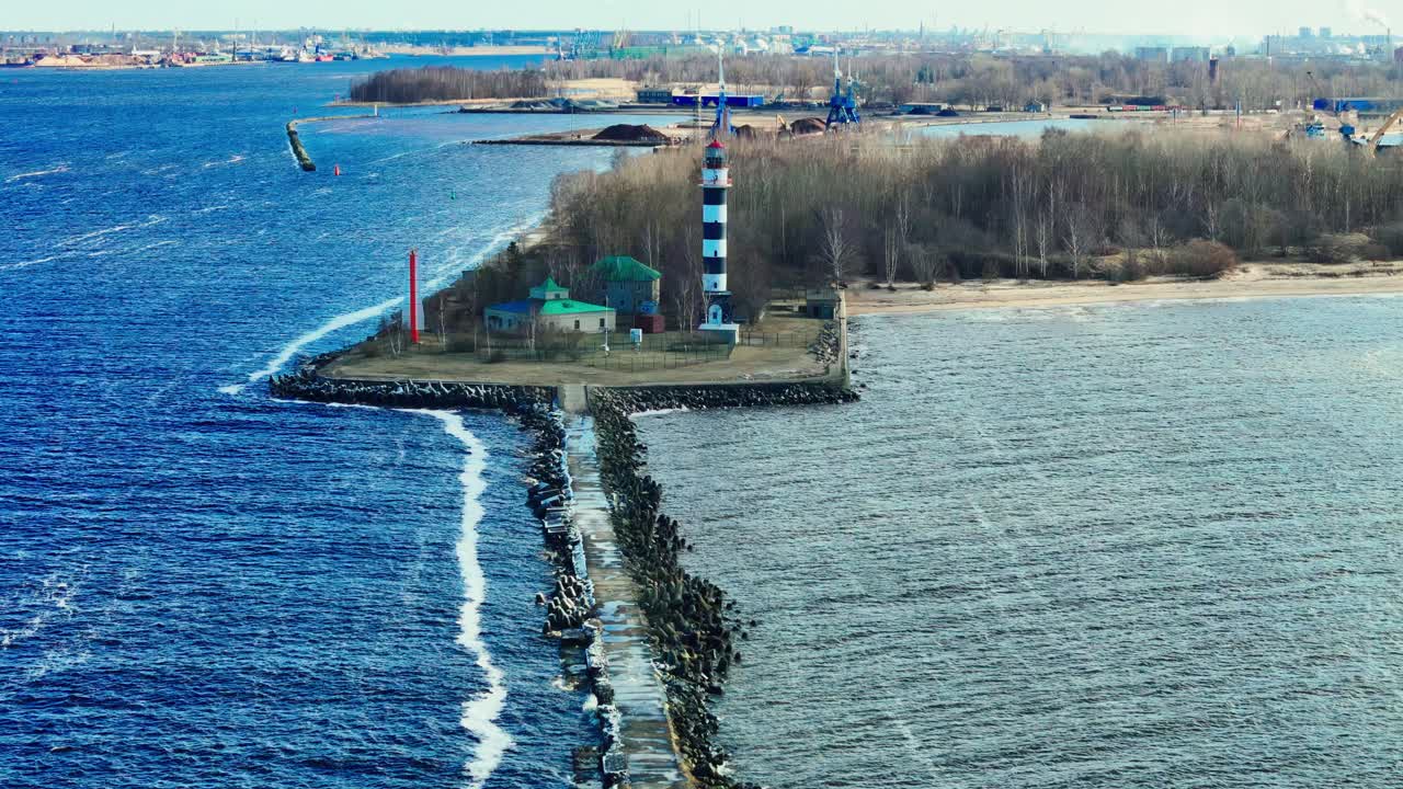 A solitary lighthouse with bold stripes rises on a stone pier jutting into cold blue waters, framed by leafless coastal trees and an empty sandy shore.