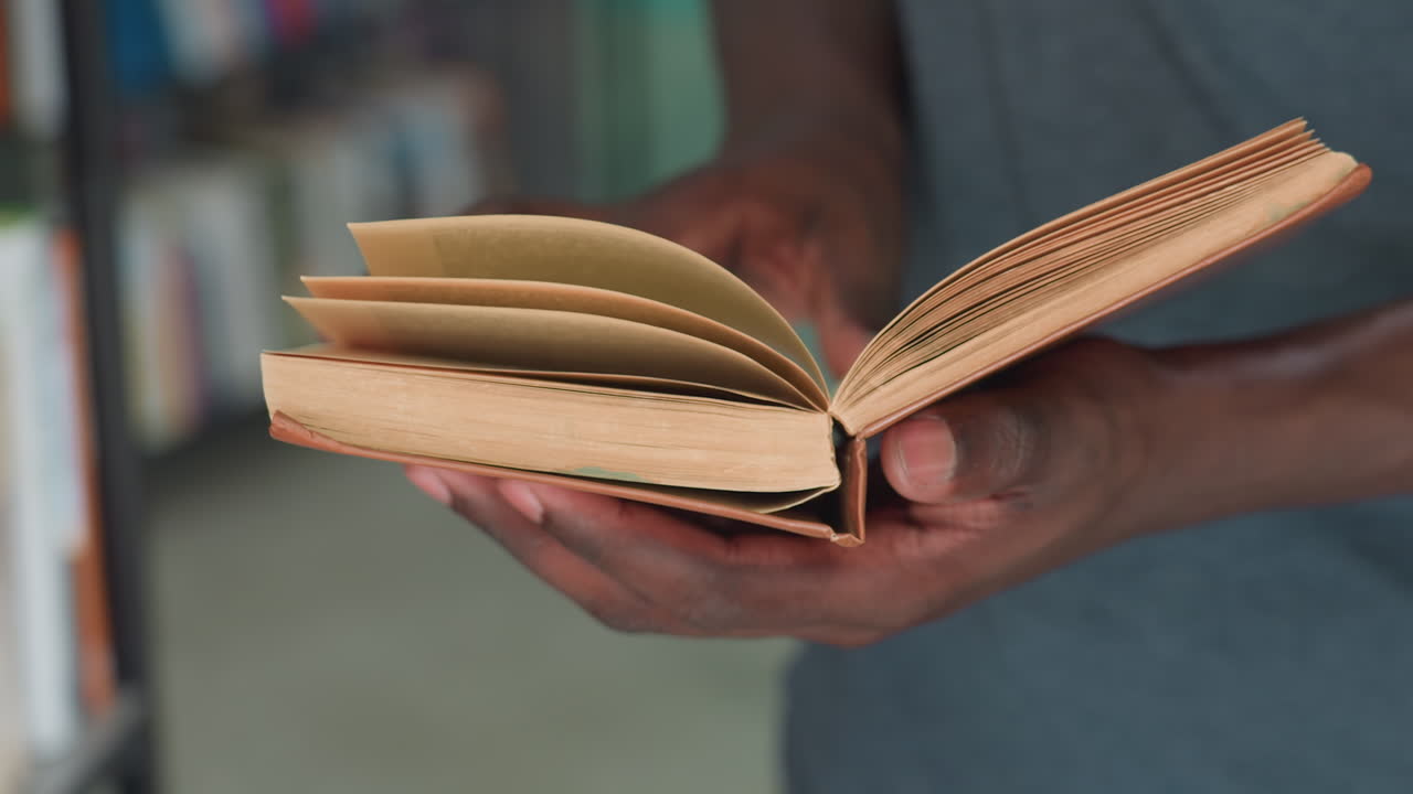Close up of dark skinned person holding open vintage book with worn pages, gently flipping through as they read, with blurred background of library shelves