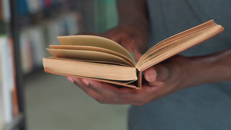 close up of dark skinned person holding open vintage book with worn pages, gently flipping through as they read, with blurred background of library shelves
