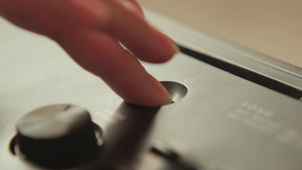 Fingertuppen trykker på en sirkulær fordypning på et elektronisk kontrollpanel ved siden av en dreieknapp, som viser taktil kontakt, en liten fordypning, varmt beigefarget lys, grunn dybdeskarphet og raffinert overflatedetalj