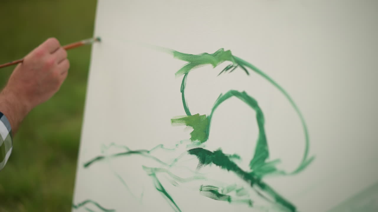 A close-up shot of a painter's hand as they create sweeping green brush strokes on a white canvas. The focus is on the hand's movement and the vivid green paint against the stark white background