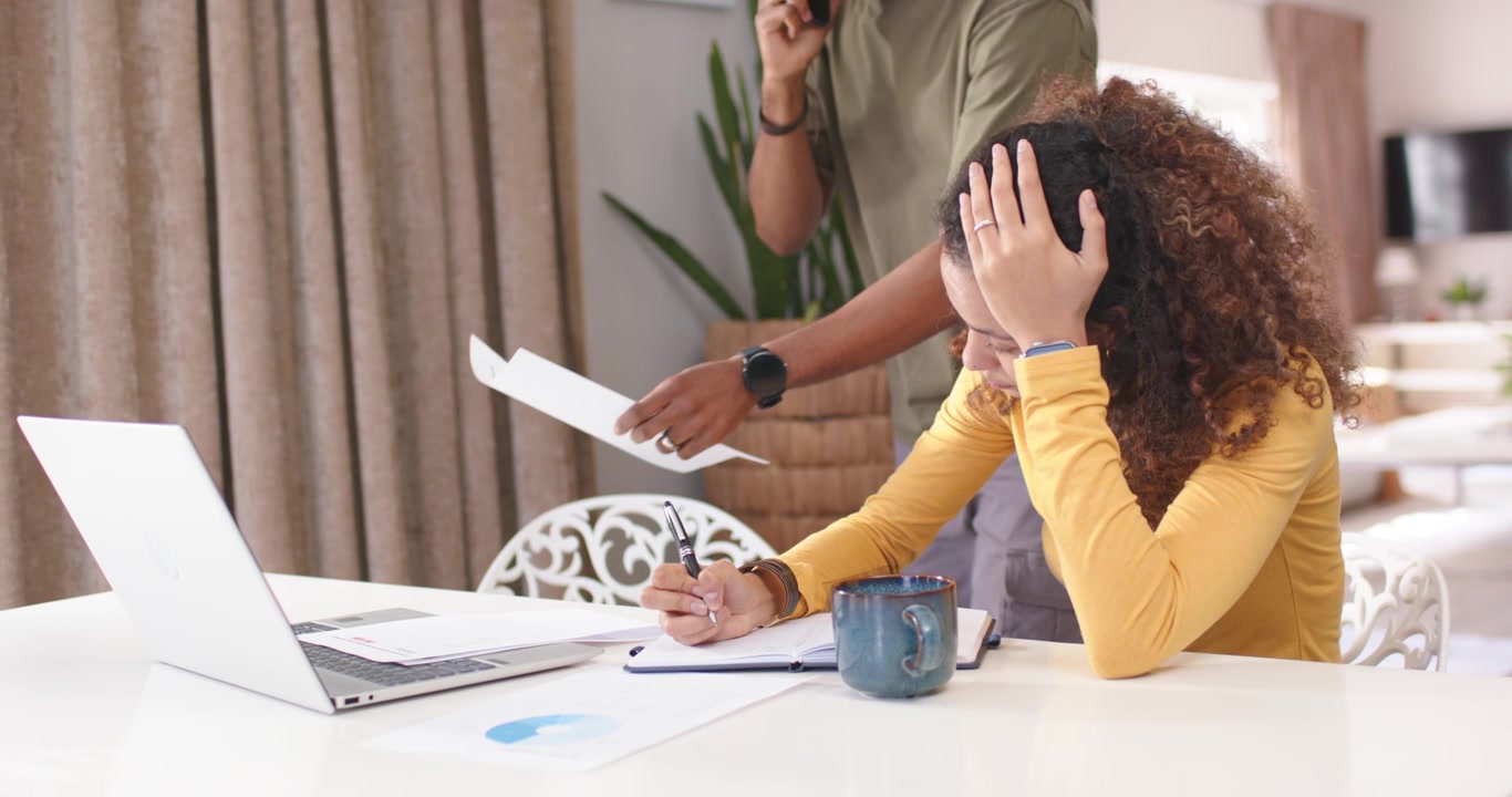 Couple at home woman in yellow writing holding head as man calling bringing papers reviewing laptop