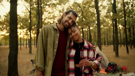 una pareja feliz, un hombre moreno con una camisa verde y una chica de piel negra con una camiseta a cuadros se paran y miran a la cámara y sonríen contra el fondo de los accesorios que llevaron con ellos en una caminata en un bosque verde soleado de verano