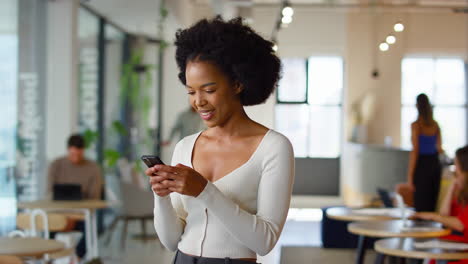 Smiling-Businesswoman-Standing-In-Busy-Open-Plan-Office-Messaging-On-Mobile-Phone