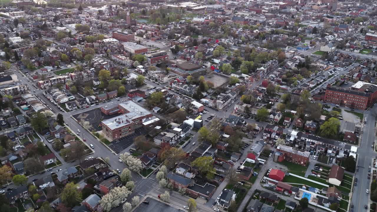 Sorvolo aereo di una storica città americana al crepuscolo in primavera, che mostra tetti colorati, strade di quartiere e classici edifici in mattoni con alberi in fiore attraverso il paesaggio urbano