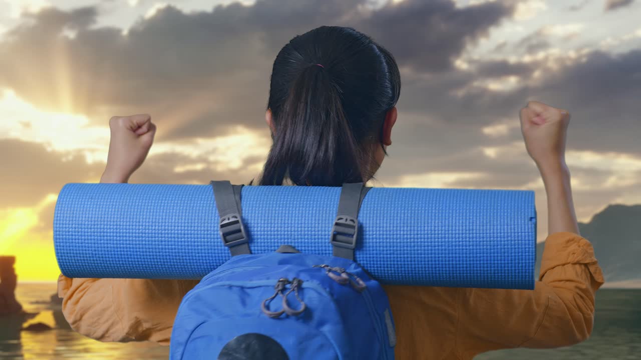 夕焼け時に浜辺に立ち、成功を祝って叫び、目標を達成したことを喜ぶ登山用バックパックを背負った女性ハイカーの後ろからのクローズアップビュー