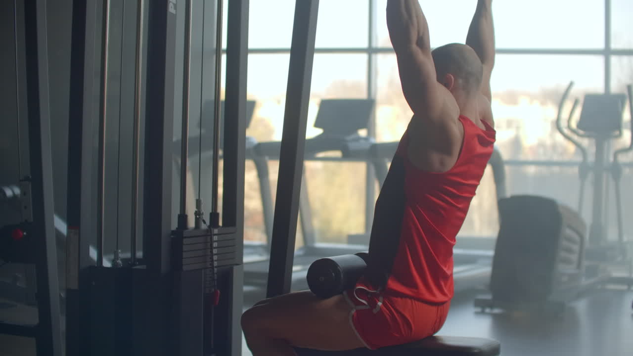 atleta haciendo ejercicios en el simulador con pesas y tira de la barra sentado en el fondo de un gran gimnasio de ventana. fuerte atleta con propósito vista desde la parte de atrás