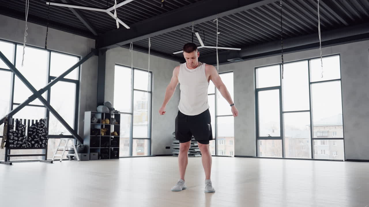 Body built man performing stretching exercise in spacious gym room with large windows and high ceiling, wearing white tank top and black shorts, preparing muscles for intense workout