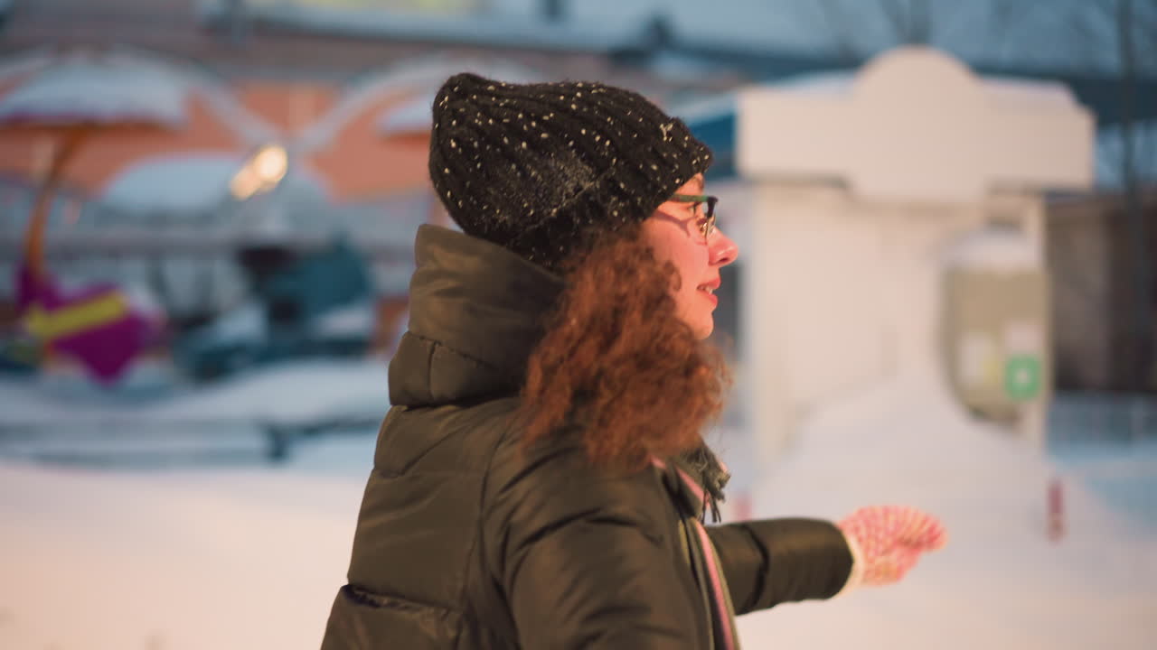 Female skater with curly hair wearing black hat, glasses, warm jacket and scarf glides outdoors on snowy evening with festive amusement park rides blurred in background, enjoying joyful winter activity