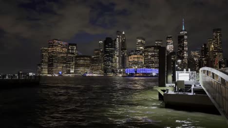 a stunning night view of lower manhattan from the waterfront, showcasing illuminated skyscrapers, the vibrant city lights reflecting on the water, and the dark, dramatic sky above