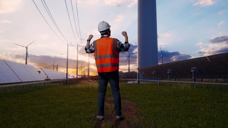 full body back view of a male engineer with safety helmet raising his hands celebrating while working with solar panel and wind turbines