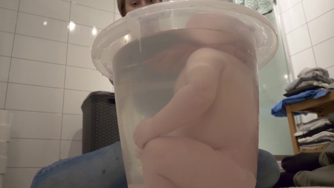 Mother cleaning newborn baby boy in clear tub sitting in bathroom. Closeup on child splashing in water.