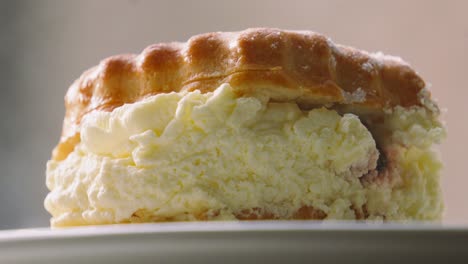 side view of sumptuous traditional cream and jam scone with light dusting of icing sugar on top resting on plate in natural light. british dessert from devon and cornwall. classic afternoon tea.