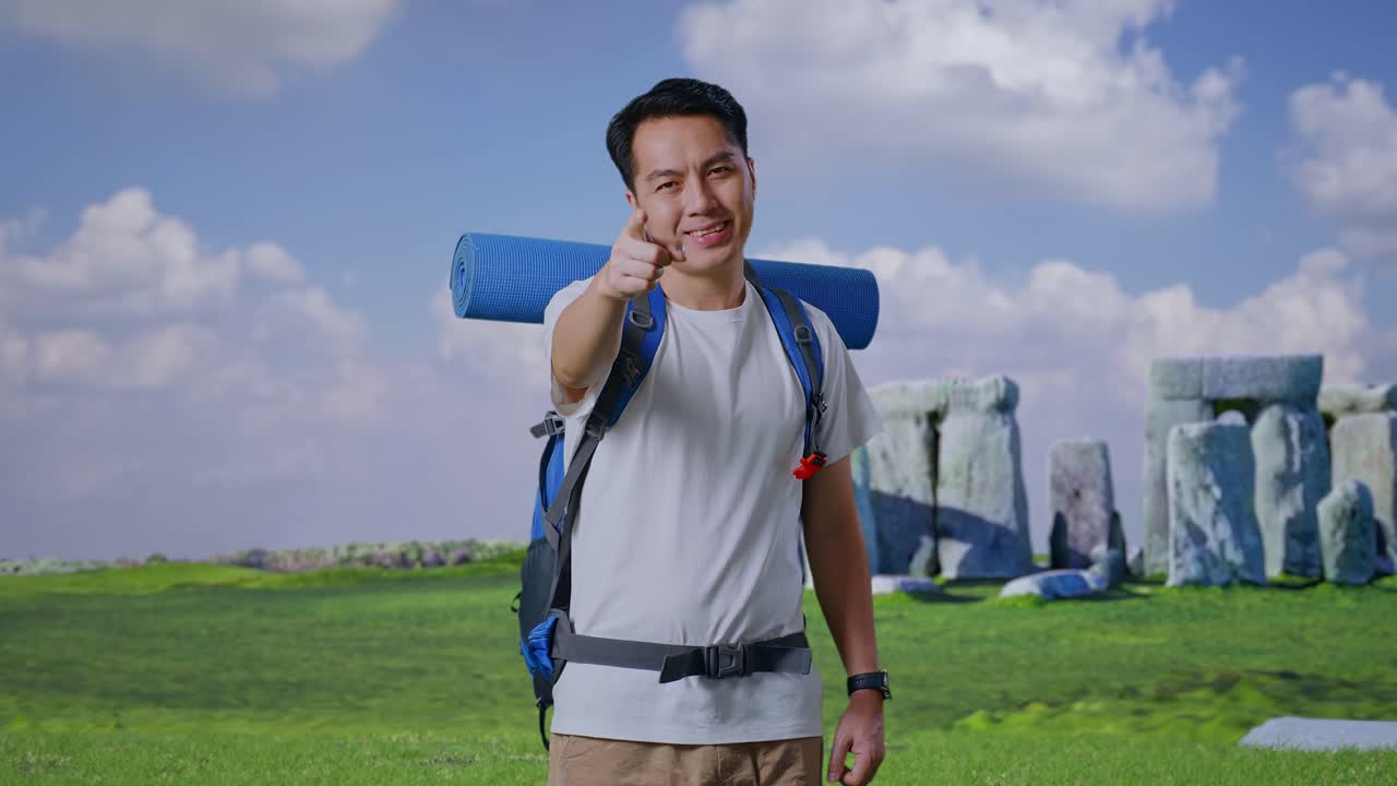 Asian Male Hiker With Mountaineering Backpack Smiling, Touching His Chest, And Pointing To Camera While Traveling In Stonehenge