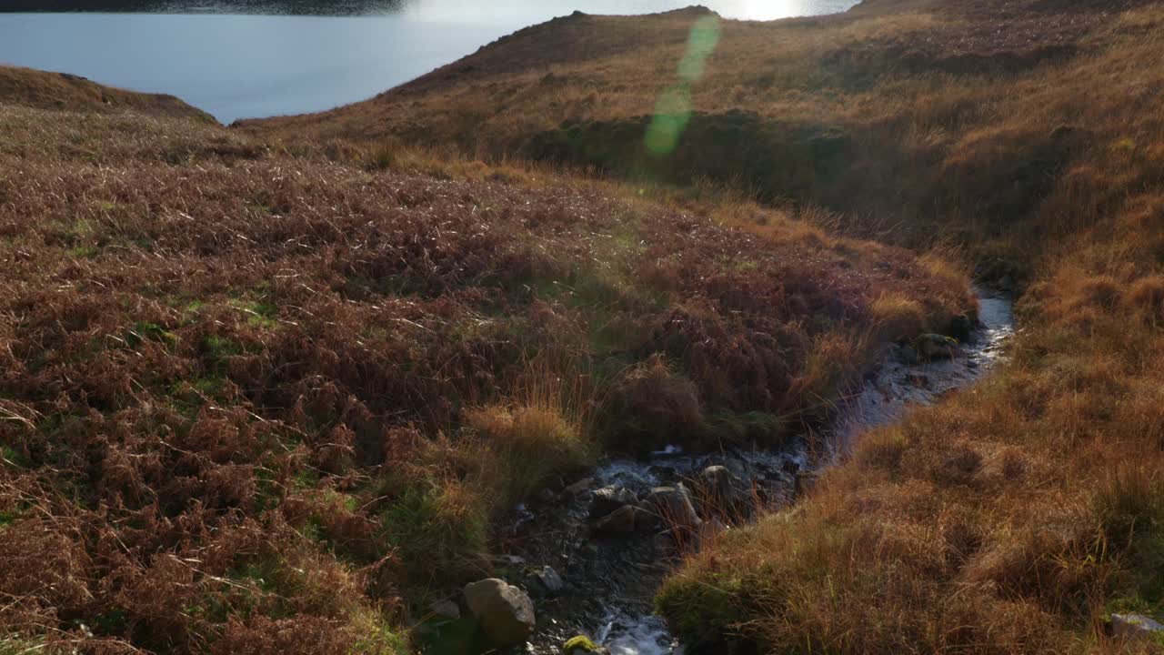 una cámara se inclina suavemente desde un río que fluye para revelar una ráfaga de sol al atardecer que se refleja en la superficie de un lago marino en las tierras altas del noroeste de escocia mientras se pone detrás de las montañas