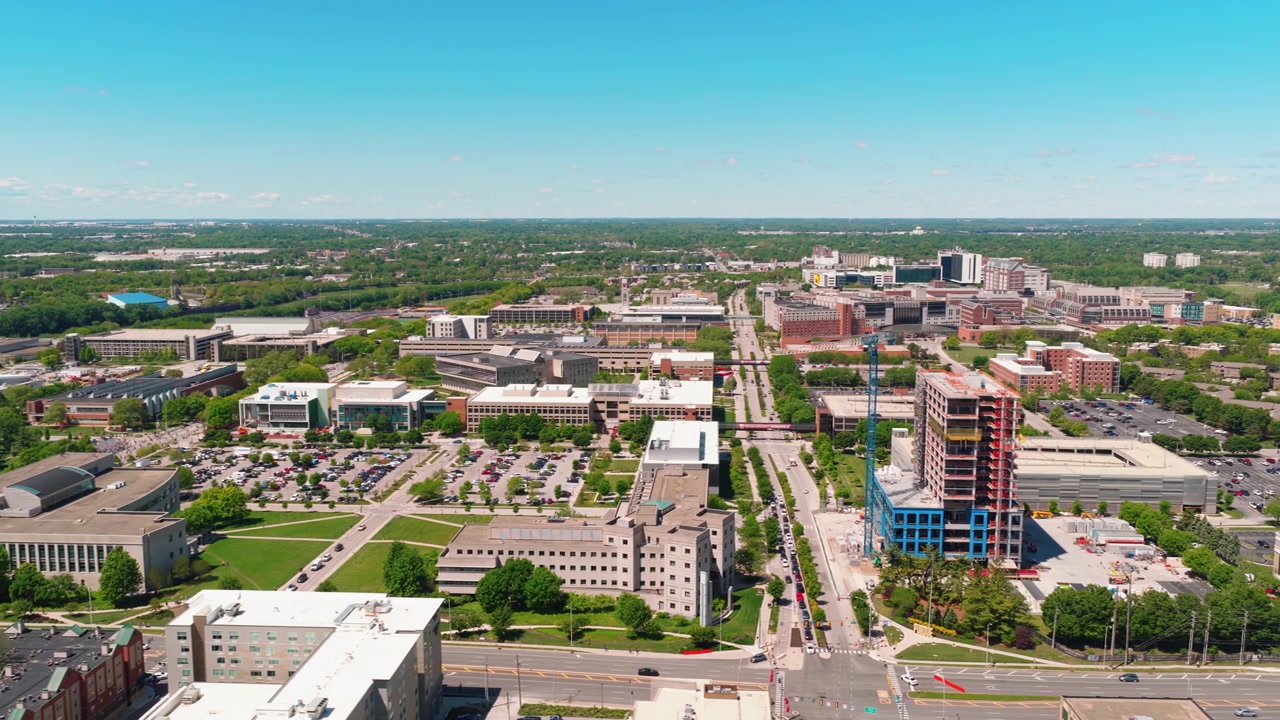 Panoramisk luftfoto av forstedene til Indianapolis som viser en blanding av kontorbygg, grøntområder og ny høyhusbygging under en klar blå himmel.