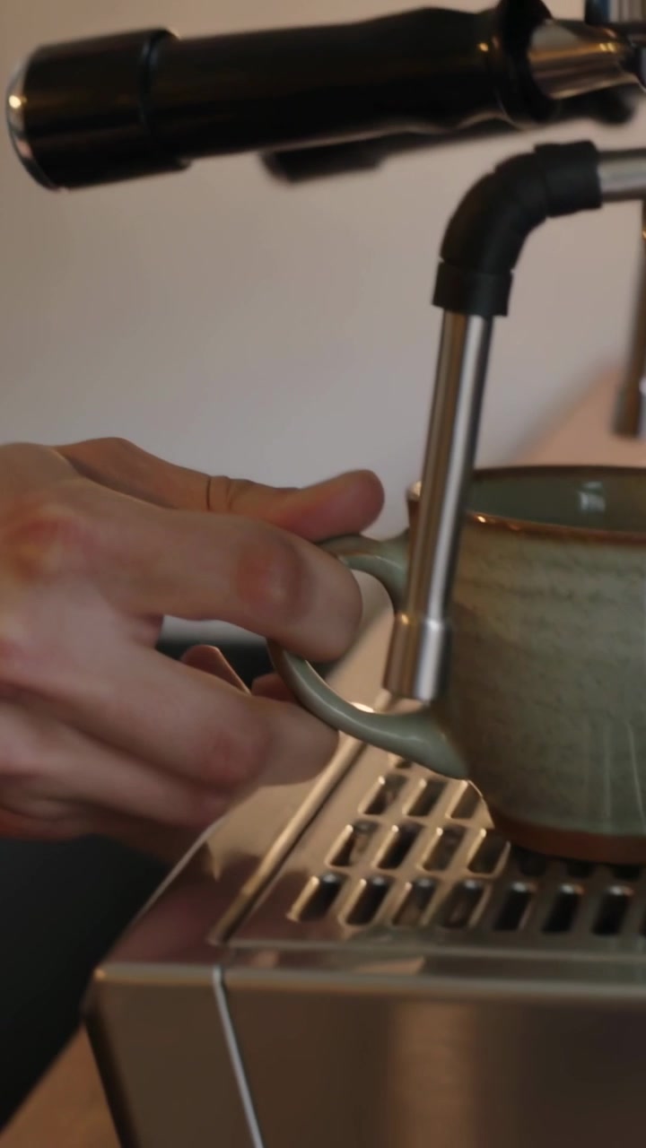 Video verticale: Uomo con grembiule di jeans che si allunga per posizionare una tazza sotto la macchina da caffè espresso che prepara il caffè in un bar.
