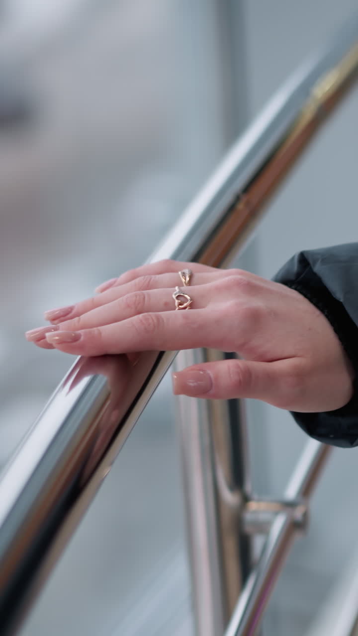 블랙 후디를 입은 여성 손의 클로즈업, 반지 장식 손가락이 매끄러운 금속 레일에서 미끄러지며, 반사 건물 유리로 부드럽고 자연스러운 빛으로 야외 도시 풍경을 보여줍니다.