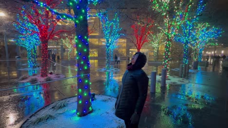 Mujer-Mayor-Atrapando-Nieve-En-Su-Boca-En-Un-Parque-De-La-Ciudad-Con-Luces-Navideñas