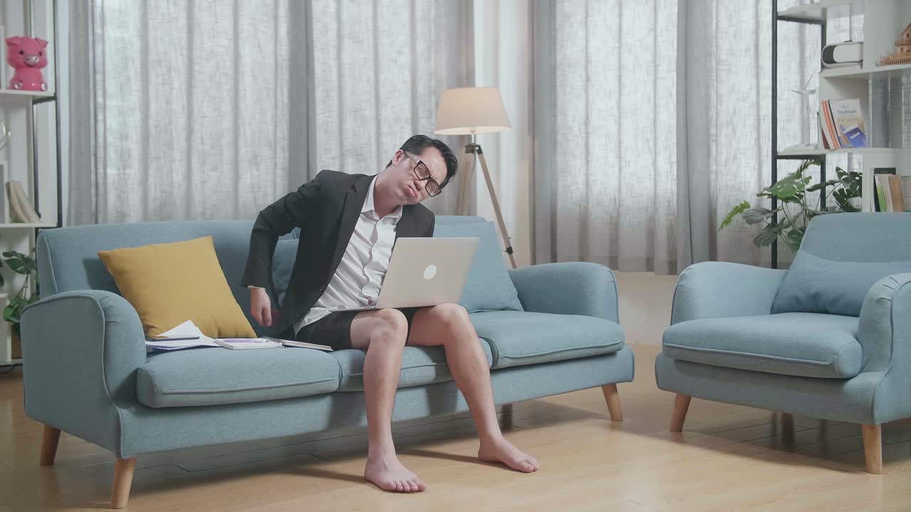 vista completa de un hombre de negocios asiático en chaqueta y pantalones cortos con dolor de espalda mientras trabaja con una computadora portátil en casa