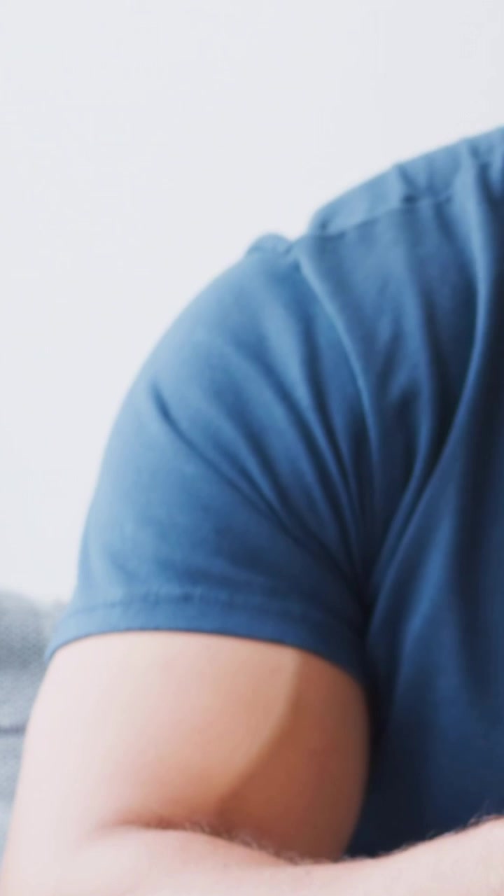 Vertical video: Shifting weight, man in blue tee turning and resting hand with braided band on sofa