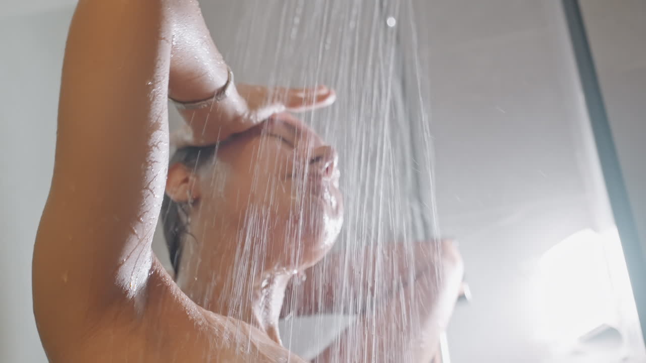 Huésped de spa en ducha de lluvia de hotel saboreando un chorro cálido, rociador de techo vierte agua constante, ojos cerrados, inhala y exhala, expresión tranquila, toalla y botellas de lujo en nicho, momento de limpieza consciente.