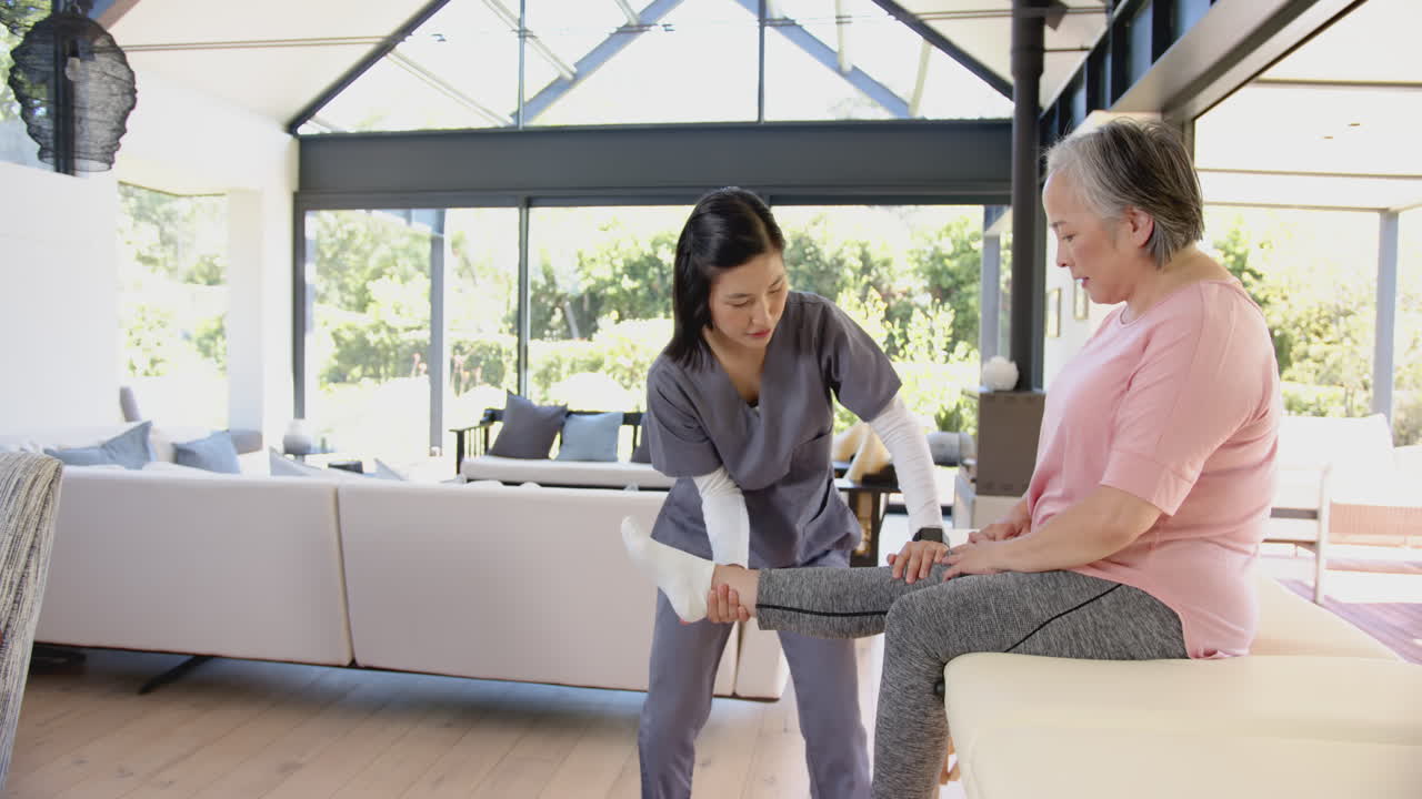 Physiotherapist assisting senior asian woman with leg exercises in home setting