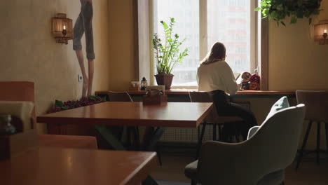 back view of lady seated by sunlit window reading book in peaceful cafe interior, surrounded by indoor plant, cozy furniture, and wall decor as soft natural light fills space with calm atmosphere