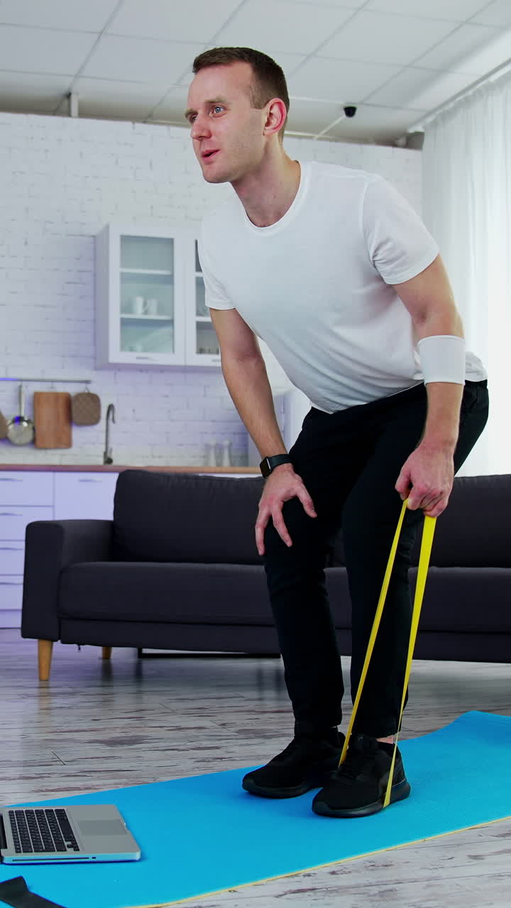 Young man training at home with a sport rubber band. Guy exercising on a mat standing in front of a laptop in kitchen. Healthy lifestyle.. Vertical video