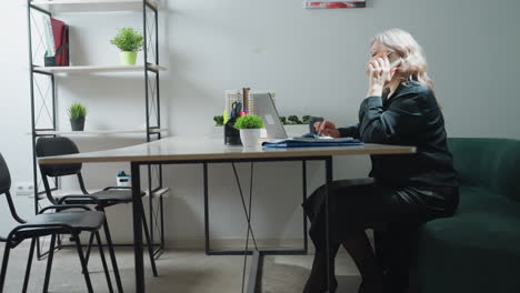 sales woman seated in stylish office multitasks by answering customer phone call while working on laptop, surrounded by organized desk items, real estate model