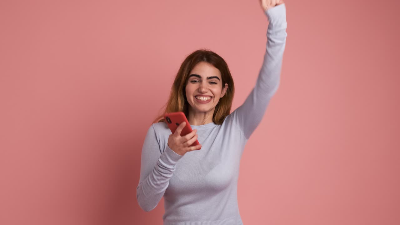 Surprised young redhead lady using mobile phone in pink background