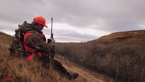 male hunter in bright orange vest and cap sitting on sidehill fiddling in his pockets and backpack