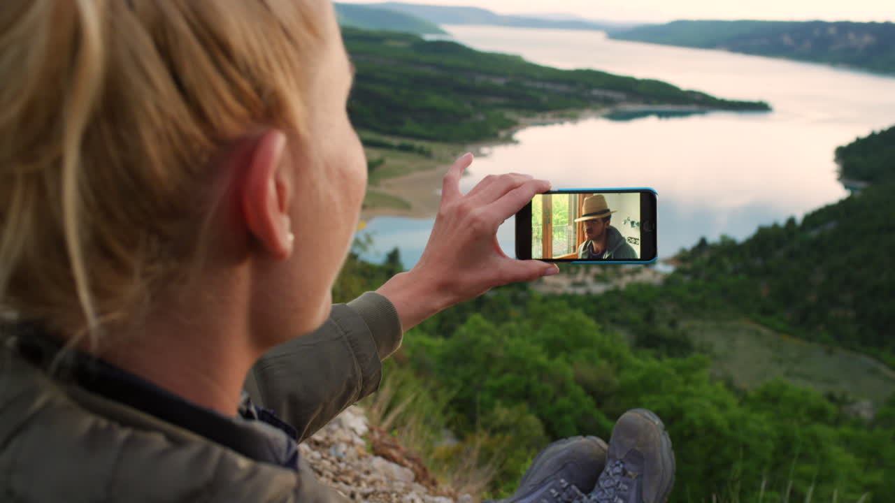 mujer hablando en video con un amigo desde la cima de una montaña con vistas a un lago