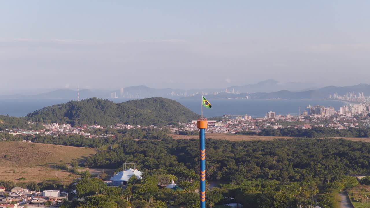 Close-up aerial footage near Beto Carrero’s striped drop tower crowned with waving Brazilian flag and backed by lush hills and distant ocean-city views. Ideal for patriotic or tourism campaigns