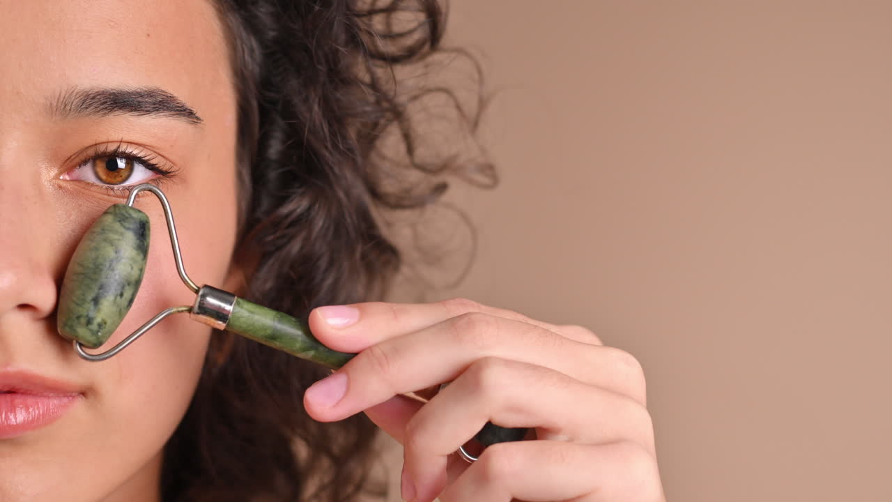 A young caucasian smiling woman is doing a facial massage using a roller, looking into the camera. Half of the face