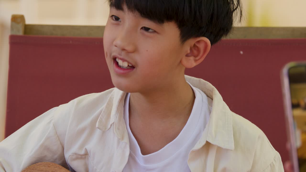 Primo piano di un ragazzo thailandese concentrato che suona l'ukulele e canta durante una lezione di musica all'aperto.