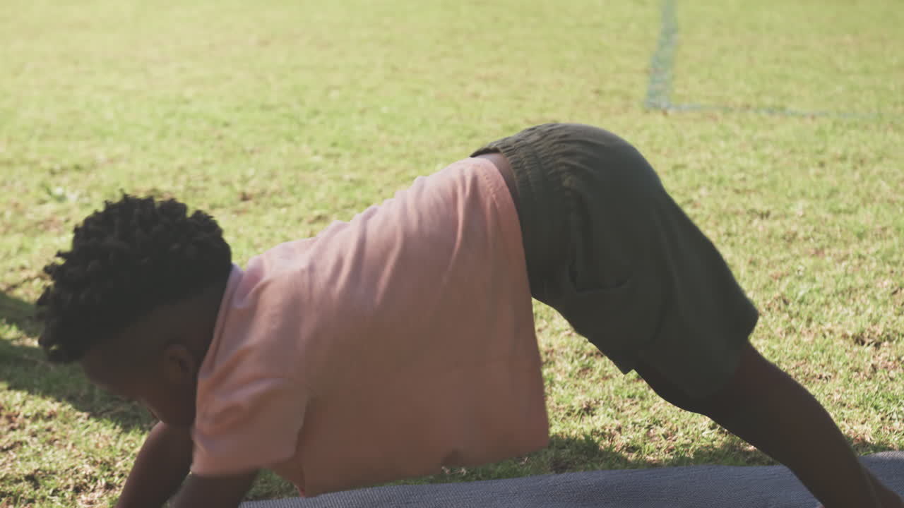 Doing yoga on mat, African American boy practicing outdoor exercise at school field