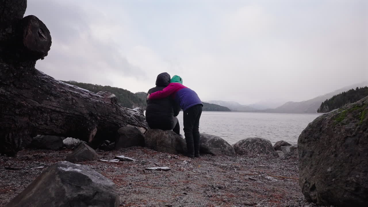 Slow motion shot of a child warmly embracing an adult seated on lakeside rocks, during snowy weather. Mist and low clouds veil the mountains across the cold lake