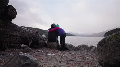 slow motion shot of a child warmly embracing an adult seated on lakeside rocks, during snowy weather. mist and low clouds veil the mountains across the cold lake