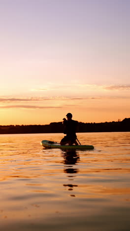 silhouette of a man wearing a cap standing on his knees on the sup board. paddle boarding on the river at sunset. low angle view. vertical video.
