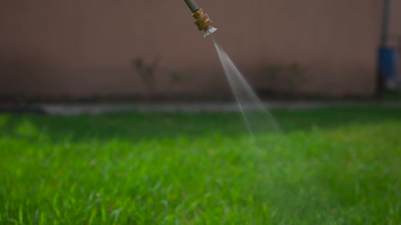 Primo piano di un ugello dell'irrigatore che spruzza acqua su erba verde brillante in un giardino domestico, con una morbida sfocatura dello sfondo e la luce naturale del giorno che evidenzia l'azione.