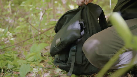 soldier places band and glove inside black bag resting on grass and carries the heavy back on the shoulder, leaving area with view of dried branches and surrounding grasses, preparing to move forward