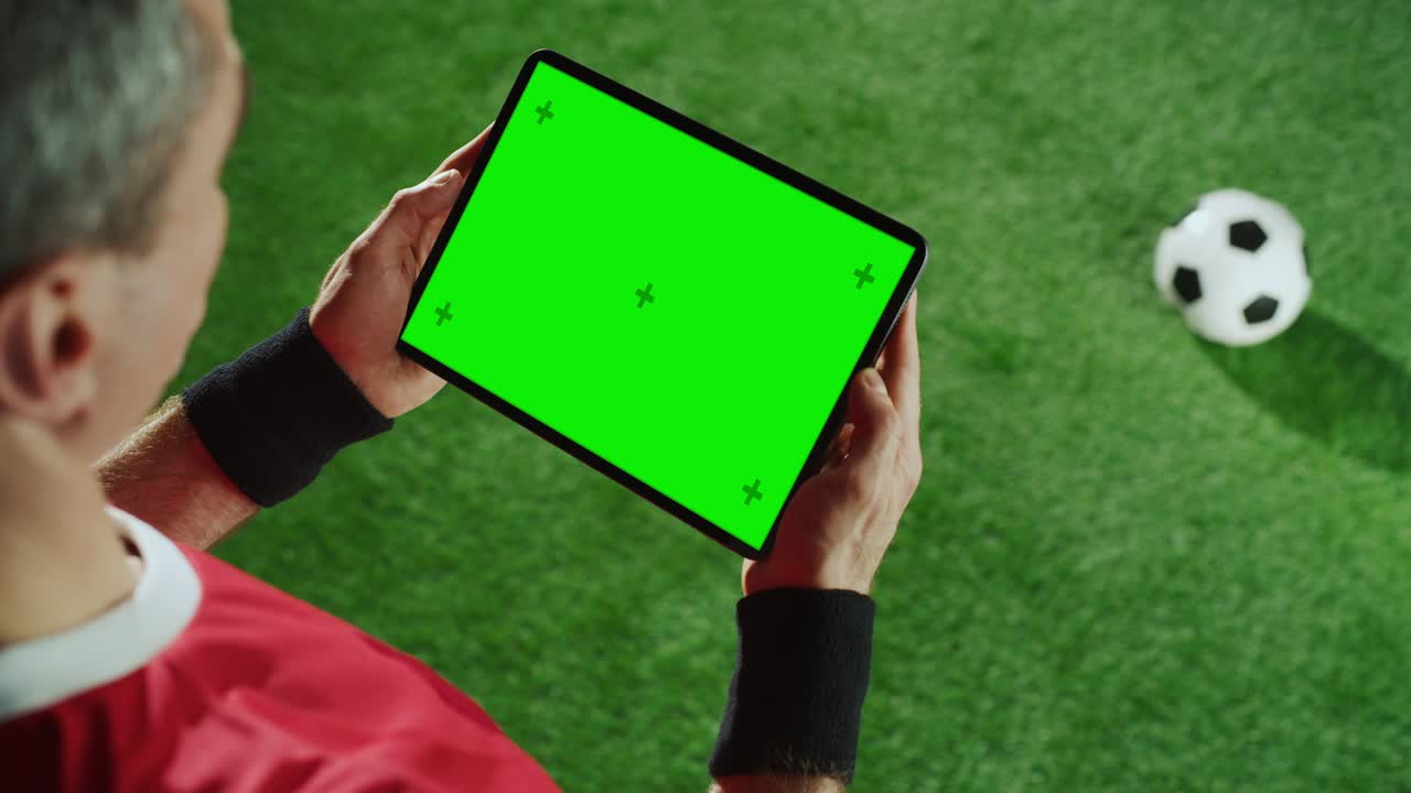 jugador de fútbol profesional sosteniendo una tableta digital con pantalla verde clave de croma con marcadores de movimiento. disparo elevado de arriba hacia abajo sobre el hombro de una estrella de fútbol usando internet, de pie en el campo