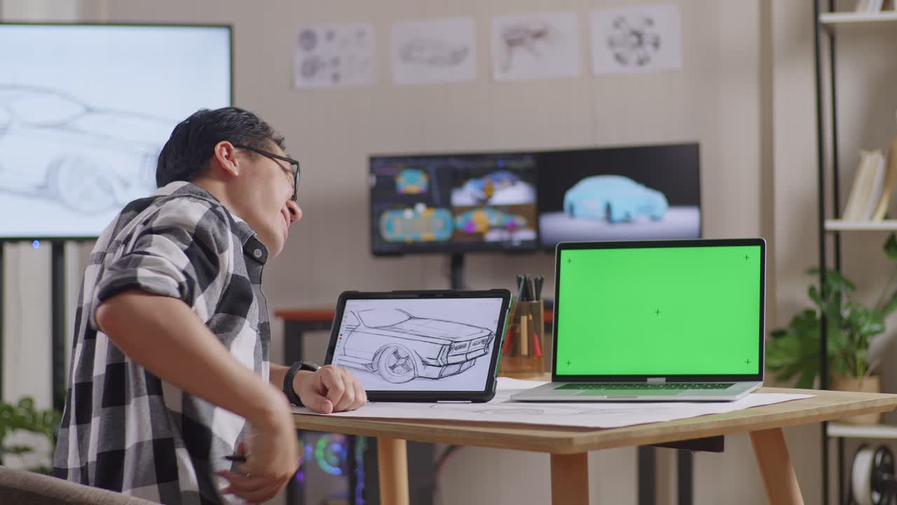테이블릿에 자동차 컨셉을 그리는 동안 허리 통증이있는 아시아 남성의 측면 뷰, tv와 컴퓨터 디스플레이 3d 전기 자동차 모델과 함께 스튜디오에서 녹색 화면 노트북 에