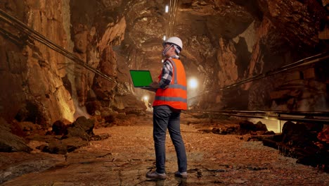 full body back view of asian male engineer with safety helmet working on a green screen laptop and looking around while standing in underground mine tunnel