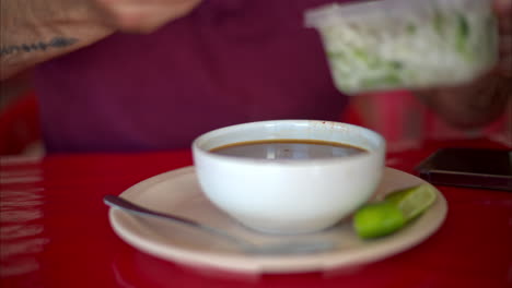 Zeitlupe-Nahaufnahme-Eines-Lateinischen-Mannes,-Der-Seiner-Barbacoa-Brühe-In-Einem-Restaurant-In-Mexiko-Gehackte-Zwiebeln-Und-Koriander-Hinzufügt