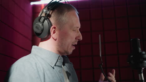 un primer plano de un hombre en un estudio de grabación con una camisa de manga larga azul y auriculares negros, cantando apasionadamente con los ojos cerrados frente a un micrófono, con un fondo de panel acústico rojo