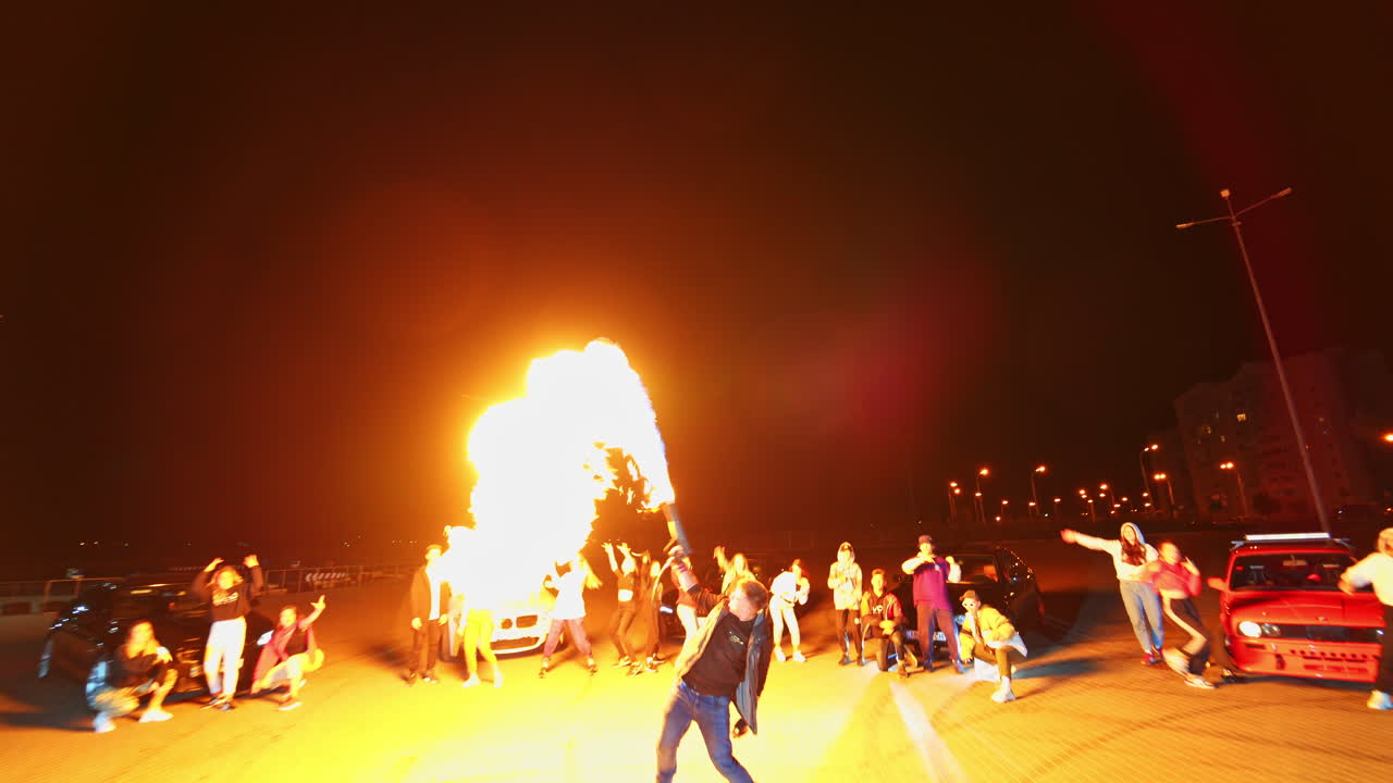 Young man performing the fire show from a special gun. Public at backdrop dances rejoicing near the cars. Night square at backdrop. Low angle view.