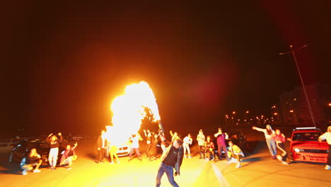 young man performing the fire show from a special gun. public at backdrop dances rejoicing near the cars. night square at backdrop. low angle view.