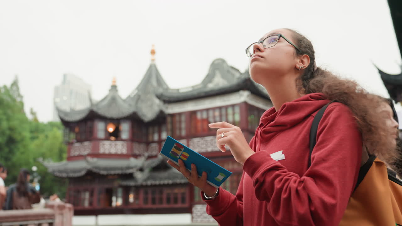 Giovane turista con felpa rossa e zaino, legge una guida vicino a un tempio. Si ferma per studiare la mappa e ammirare l'architettura ornata della pagoda, in un momento spontaneo di esplorazione urbana, con calma curiosità per il patrimonio culturale.